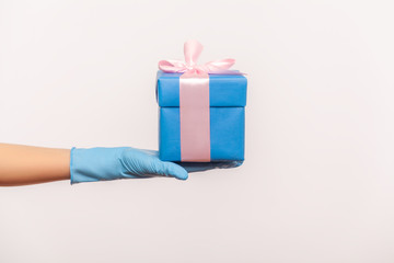 Profile side view closeup of human hand in blue surgical gloves holding blue gift box. sharing, giving or delivery concept. indoor, studio shot, isolated on gray background.