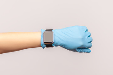 Profile side view closeup of human hand in blue surgical gloves holding and showing wirst smart watch screen. indoor, studio shot, isolated on gray background.