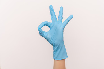 Profile side view closeup of human hand in blue surgical gloves showing Ok sign or number 3 with fingers. indoor, studio shot, isolated on gray background.