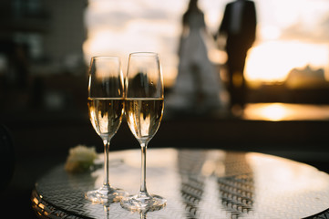 Two glasses of champagne stand on a table stand on a table. A loving couple of newlyweds is standing in the background. 