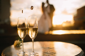 Two glasses of champagne stand on a table stand on a table. A loving couple of newlyweds is standing in the background. Wedding rings fall into glasses