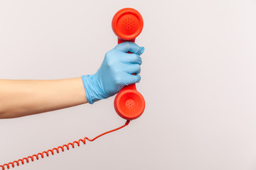 Profile side view closeup of human hand in blue surgical gloves holding and showing red cale telephone handset receiver. indoor, studio shot, isolated on gray background.