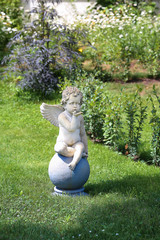 Statue of cute angel air kissing and sitting on the ball on the green background of summer lawn....