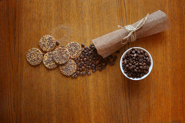 A handful of coffee beans in the small cup, some biscuits, canvas napkin on the wooden backdrop, top view.  Coffee beans scattered on the wooden table with cookies and folded napkin. Copy space