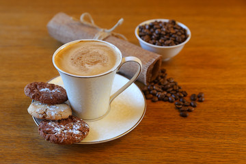 A cup of coffee with buscuits on the wooden table with coffee beans, canvas napkin, without tablecloth. A cup of cappuccino with tasty biscuits on the white saucer closeup. Wooden table. Copy space