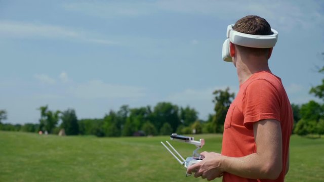 Man controls a quadrocopter through a remote control and looks at the video with goggles on his head. Drone operator in a red T-shirt and blue shorts in park is filming drone video