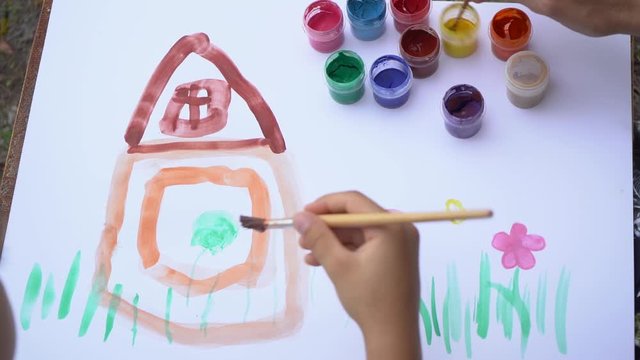 Closeup view video footage of little boy of 5 years old painting outdoors together with his mommy. Child paints cute funny house and colorful flowers on white sheet of paper.
