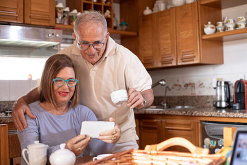 Coppia di anziani divertiti in cucina fanno una video chiamata con lo smartphone mentre fa...