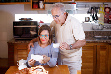 Coppia di anziani divertiti in cucina fanno una video chiamata con lo smartphone mentre fa...