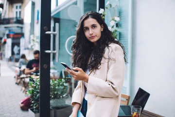 Half length portrait of millennial female user with modern cellphone device in hand posing at street urbanity, beautiful smart casual hipster girl with digital smartphone looking at camera outdoors