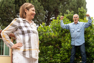 coppia di anziani in giardino gioca simpaticamente: l'uomo esulta sopra la catasta di legno e la moglie ride allegramente - obrazy, fototapety, plakaty