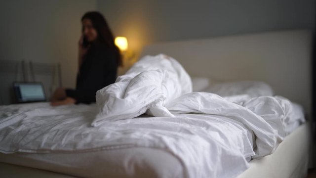 Woman sitting on bed working on laptop and mobile phone in bedroom, unmade bed with crumpled bed sheet, blanket and pillows after sleep waking up in the morning.