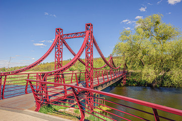 beautiful iron red suspension pedestrian bridge over the small river Irpin from the promenade to the green forest.