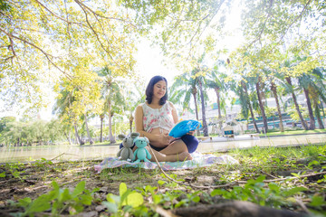 Naklejka na ściany i meble Portrait of happy pregnant asian woman looking at dool sitting in a park at sunrise