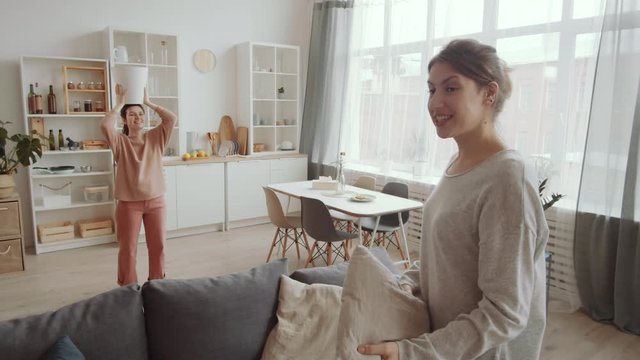 Female roommates having fun in isolation at home: young woman waving at camera and greeting viewer, then throwing pillow to her friend while she standing with trash bin on head but missing can