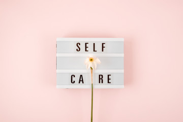 Self-care word on lightbox and flower narcissus on pink background flat lay. Take care of yourself.