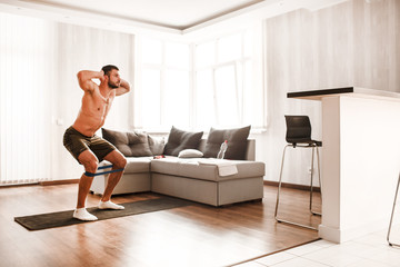 Obraz premium T-shirtless guy sportsman in go in for sport at home. Young man doing sport workout in room during quarantine. Stand on yoga mat and stretch leg on back using resistance band doing squats.
