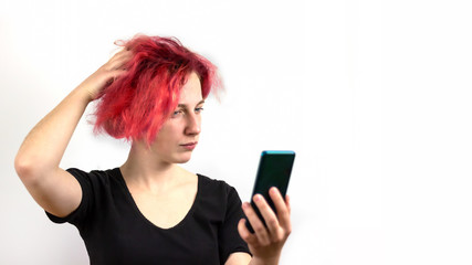 A rocker girl with red hair is talking on a video link on a smartphone, her hand is pulling her hair, on a white background. Concept of informal youth