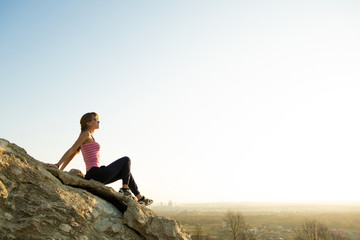 Fototapeta na wymiar Woman hiker sitting on a steep big rock enjoying warm summer day. Young female climber resting during sports activity in nature. Active recreation in nature concept.