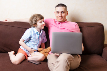 Surprised young man sit on computer using laptop relax with preschooler son have fun together, smiling dad and little boy child enjoy weekend at home rest on sofa busy with gadgets. Online education.