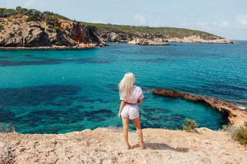 Blonde sexy girl looking at Cala Portinatx from the coast in Ibiza, Spain. Beautiful woman looking to the blue aqua sea. Back view.