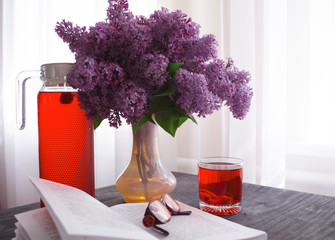 Still life with a bouquet of lilacs and compote of berries. A glass of compote, a jug, a bouquet, a book and glasses on a dark wooden table. Still life near the window.