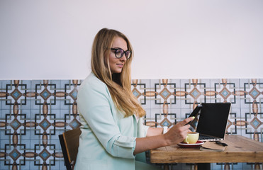 Caucasian millennial woman dressed in smart casual clothing checking received email on modern cellular phone typing text for sending sms message, skilled female using cafeteria internet connection