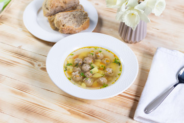 Meatball soup with potato in white plate, bread, spoon, daffodils on wooden background