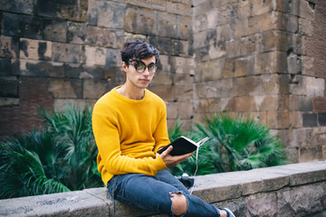 Fototapeta premium Portrait of pensive handsome male in spectacles for vision correction enjoying free time on self education, serious hipster guy looking at camera holding new book bestseller standing outdoors
