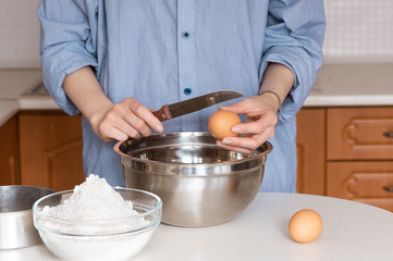 A woman prepares dough in a metal bowl: break an egg into a bowl with dough. On the table are the ingredients for the dough: flour, eggs. Cooking at home. Series.