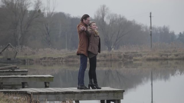 Joyful Caucasian couple standing on pier by lake with stretched hands and imitating fly. Wide shot of happy loving man and woman dating outdoors on cloudy day. Love, bonding, lifestyle, joy.