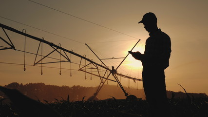 Silhouette man uses a smartphone against the background of the irrigation system in the field at sunset