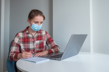 Woman in medical mask using laptop computer, watching webinar, lecture, making notes in grey room at home. Communication, education, freelance, internet, quarantine, self isolation, technology concept