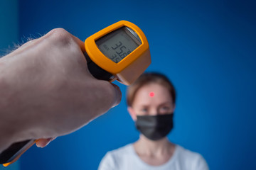 Measurement, healthcare, infection, coronavirus concept. Man hand holding yellow pyrometer and measuring temperature of woman in medical face mask looking at camera: close up view, selective focus.