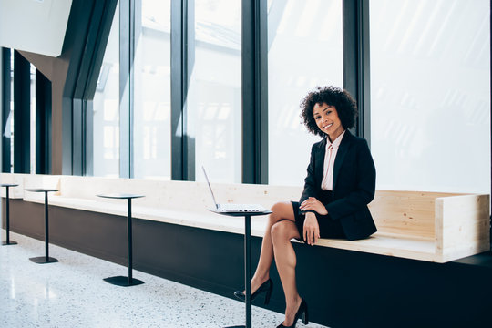 Portrait of successful afro american office worker sitting in modern workspace near table with laptop computer and smiling at camera, attractive woman dressed in elegant formal wear indoors
