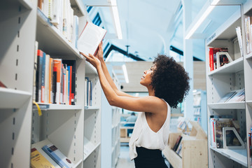 Young african american woman taking book from high shelf in library searching literature for self...