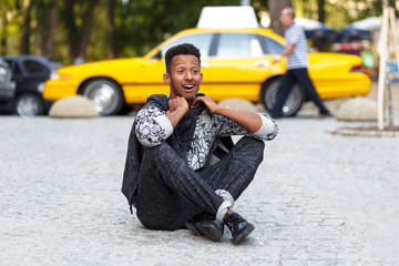 Amazed young man seated down on pavement road with crossed legs, isolated on a yellow blurred taxi background.