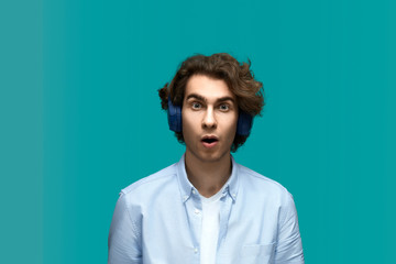 Fototapeta na wymiar Wondered of sounds. Portrait of a young beautiful man wearing white t-shirt and blue shirt in headphones surprised from music content