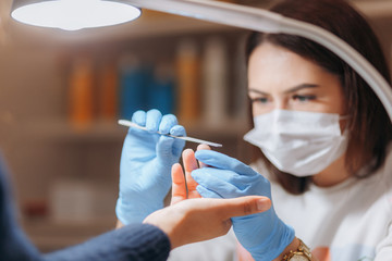The master uses a nail file to align the texts of the nail during the manicure. The master has a mask on his face