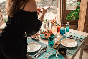 Lovely rich woman with curly hairstyle have a romantic dinner at restaurant. Overwhelming girl in black dress with colorfull cocktail. Lady eating in a restaurant