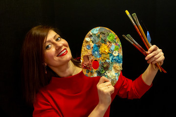Fototapeta premium A young beautiful woman wearing red dress on black background. pretty young smiling woman holding a palette and artistic brushes.