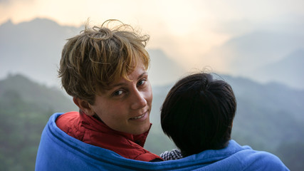 A young beautiful international couple, best friends are sharing a blanket and watching a sunrise in the mountains from aerial peak view,  smiling happy woman man, summer  vacation, travel concept