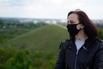 Young woman in black medical mask and leather jacket on hill on cloudy day. Adult female walking alone in countryside.