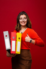 Confident girl in glasses and with folders in her hands motivates and charges for success. Isolated on red background