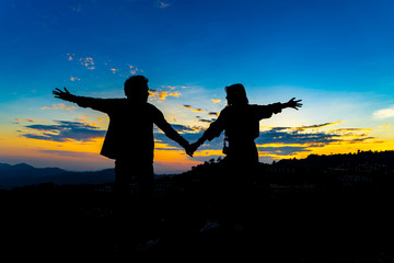 Fototapeta na wymiar Silhouette of Young Asian Couple Holding Hand at Sunset on Mountain with Blue Sky. Enjoying Peaceful Moment Concept. Relaxing or Love Family Relationship Concept.