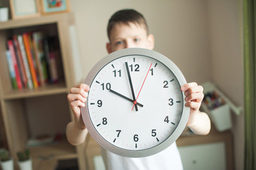 A teenager boy holds a big clock with large numbers on outstretched hands. A child in a white T-shirt, watch closeup, face in defocus. In the children's room. Concept mode, schedule, sleep.