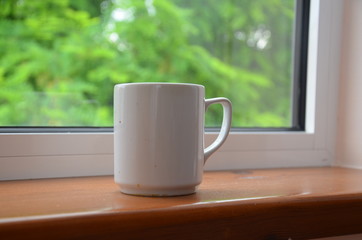 A white cup of tea or coffee stands on a wooden window sill against the background of a summer garden