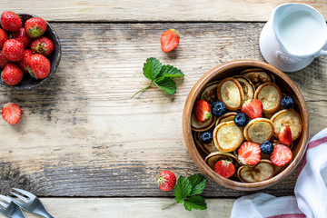 Tiny pancake cereal and chocolate mini pancakes in a wooden bowl with honey and strawberries on a wooden background. Top view. Copy space. Trendy food