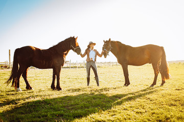 Beautiful young woman in a hat and gloves with two broun horses in a field on a sunset. Horseback...