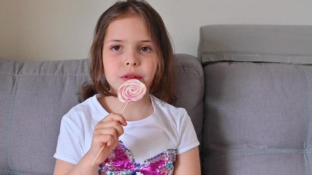 Little girl eating a big round lollipop. Rainbow candy in the hands of a child. Love and affection for sweets. A girl with blond hair 7 years old in the room.